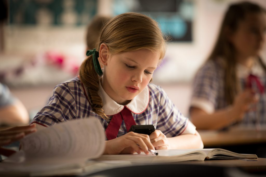 A schoolgirl in uniform reads from a book, holding a calculator