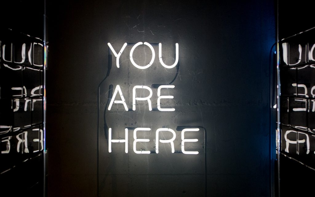 A white neon sign reads "you are here" all in upper case against a dark background. The tiled wall on either side of the sign partially reflects the words in mirror image.