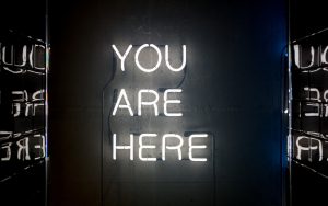 A white neon sign reads "you are here" all in upper case against a dark background.