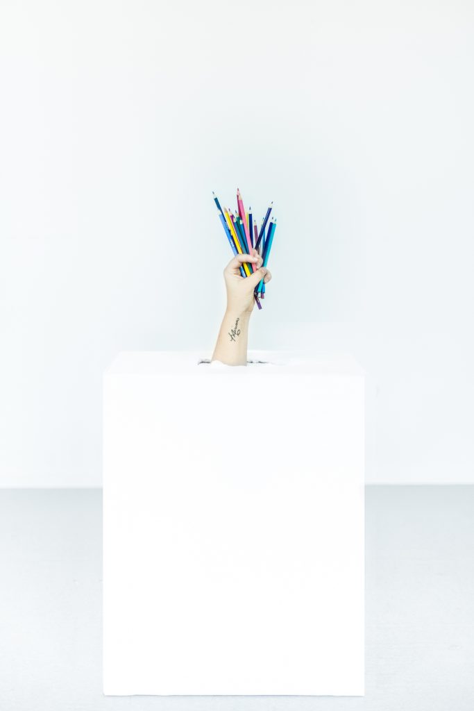 A light-skinned hand holding a bunch of colourful pencils pushes through the top of a white box, with a white background. There is a script tattoo on the wrist. 