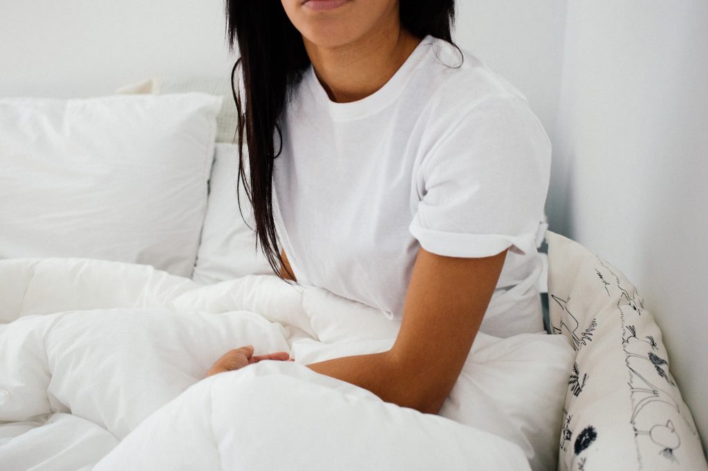 A person with long hair wearing a white t-shirt sits in bed leaning on a pillow