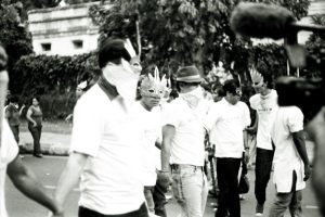 in a black and white image, a group of people march wearing feathery masks and scarves over their mouths to hide their identities.