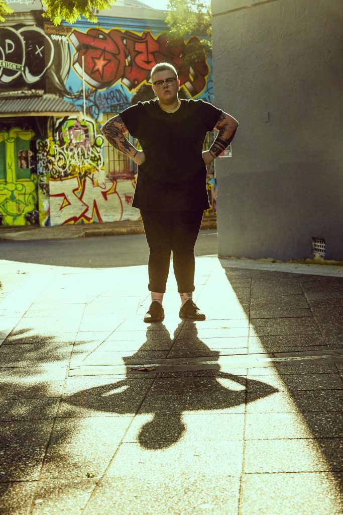 a butch person stands with their hands on their hips in front of a graffiti wall