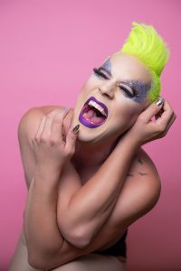 a white person with fluoro yellow hair and a full face of makeup yells at the camera, their arms covering their nude torso