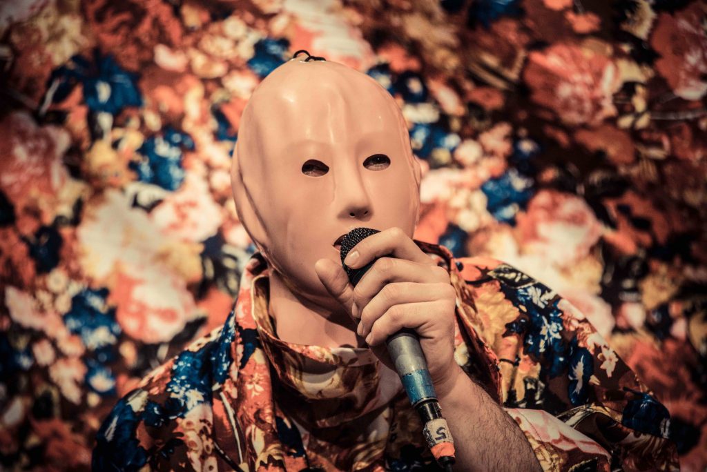 a person in a mask holds a microphone to their mouth