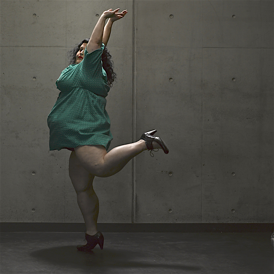 Kelli Jean wears a short green dress, and has their hands above their head with one leg kicked behind them. 