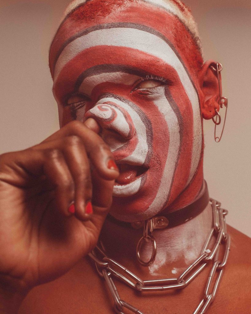 a person has their face painted with red and white circles, the whites of their eyes visible, and their hand touching their nose.