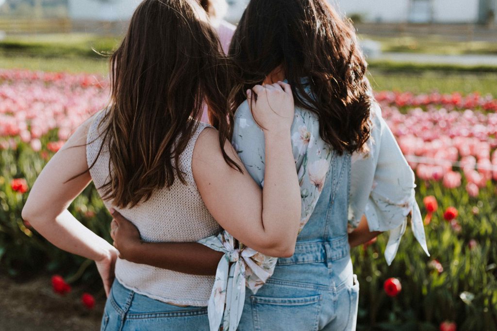 Two femme presenting people stand arm in arm in front of flowers, with their backs to the camera.