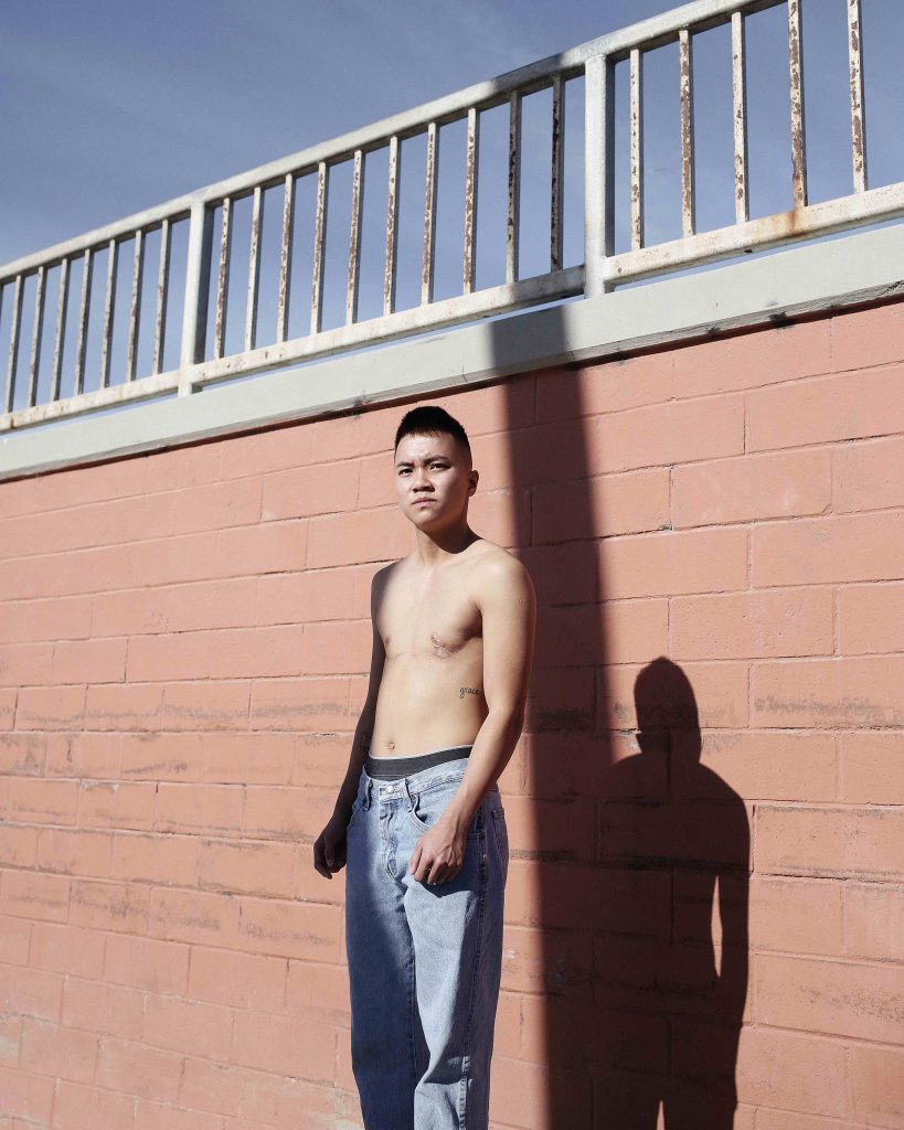 A person stands, facing the camera, shirtless, in front of a brick wall. They have scars and tattoos on their chest. 
