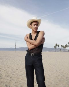A person wearing black and a cream cowboy hat wraps their arms around themselves, looking to the side, with a beach backdrop.