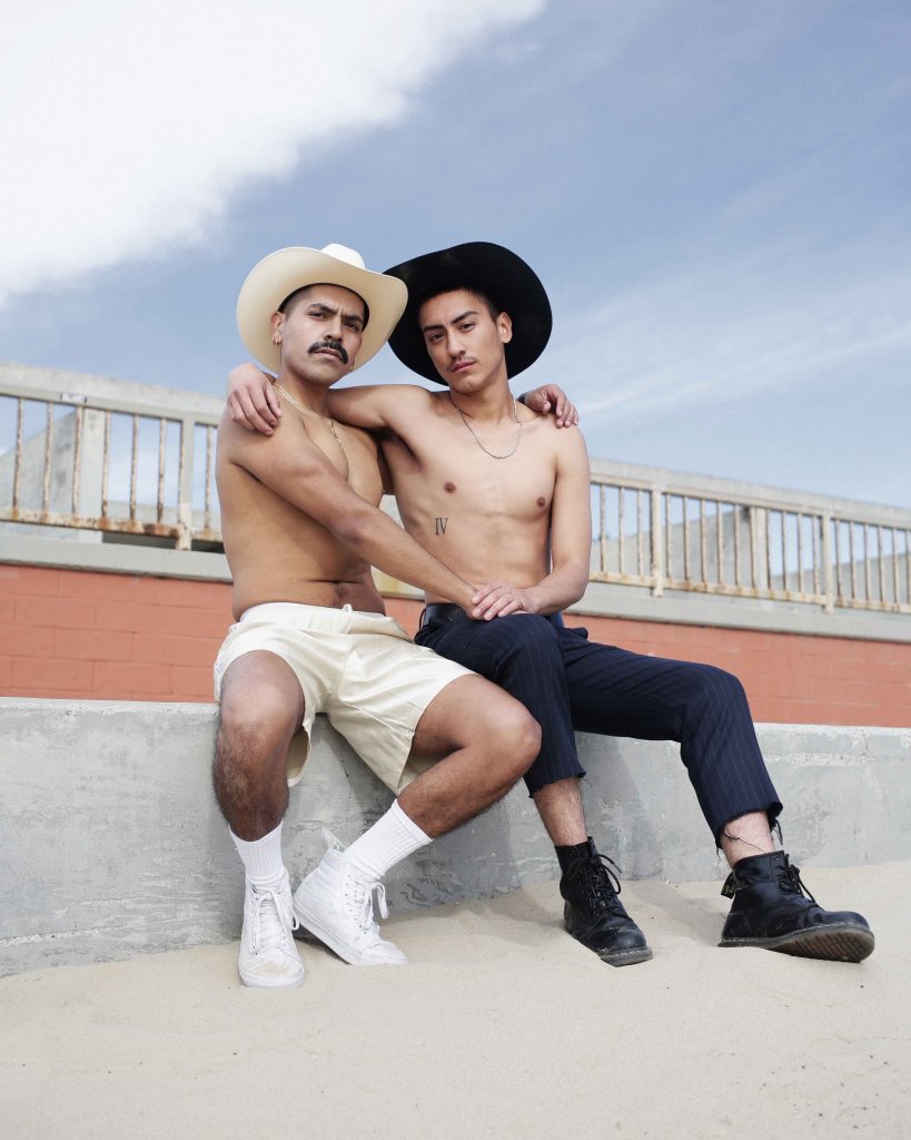 Two shirtless people sit arm in arm. One is wearing cream/gold/white, and the other is dressed in black (apart from their bare chests). 
