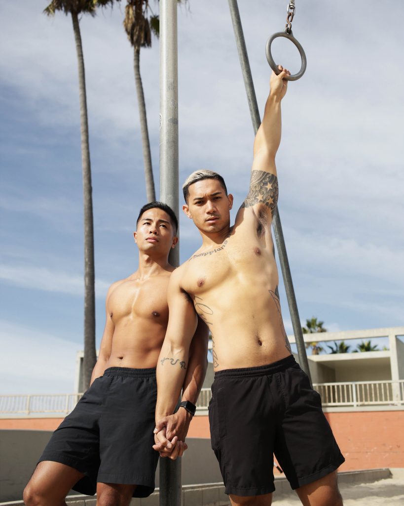 two shirtless people stand, facing the camera among outdoor gym equipment. One of them is holding a gym ring above their head. 