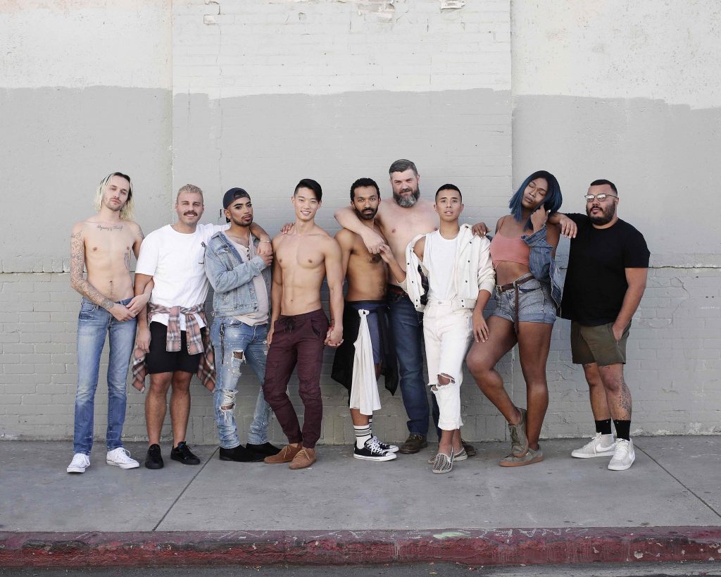 A group of people of different sizes and races stand facing the camera. Some are shirtless, some are clothed. 