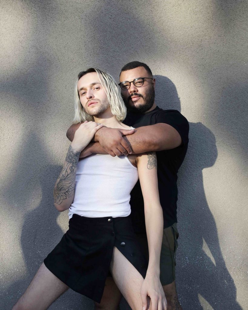 Two people stand, embracing, facing the camera. One has glasses, a beard and a black tshirt, while the other has shoulder length blonde hair, a white singlet and a black skirt. 