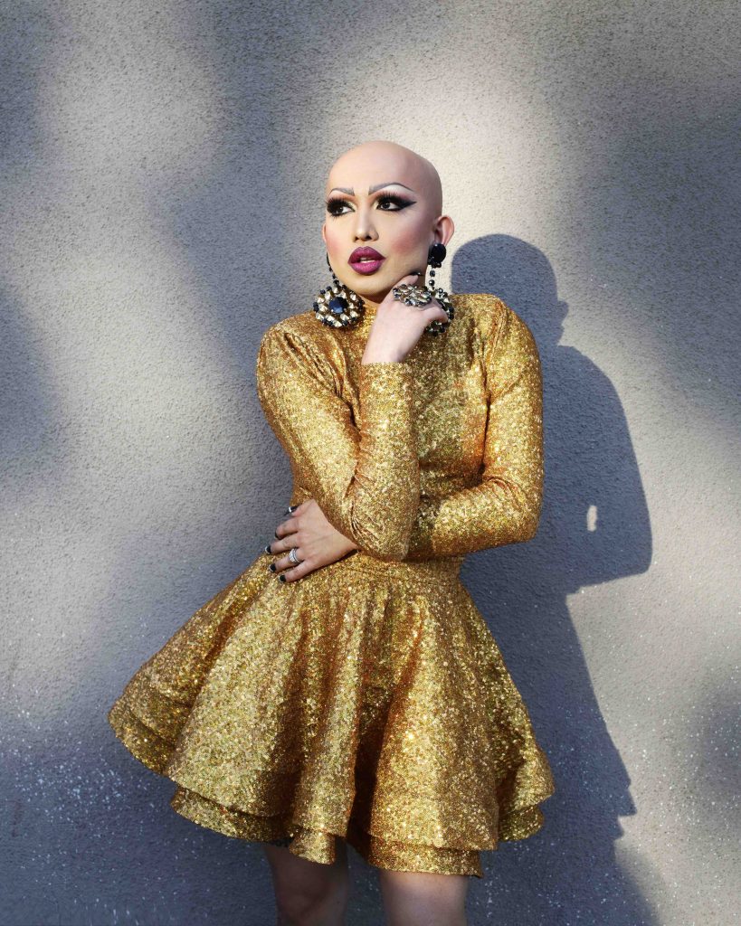 A person with no hair, a full face of makeup and a sparkly gold dress looks to the side in front of a concrete backdrop.