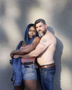 Two people stand, spooning, facing the camera. One is shirtless with a beard and tattoos, while the other has blue and black shoulder length hair.