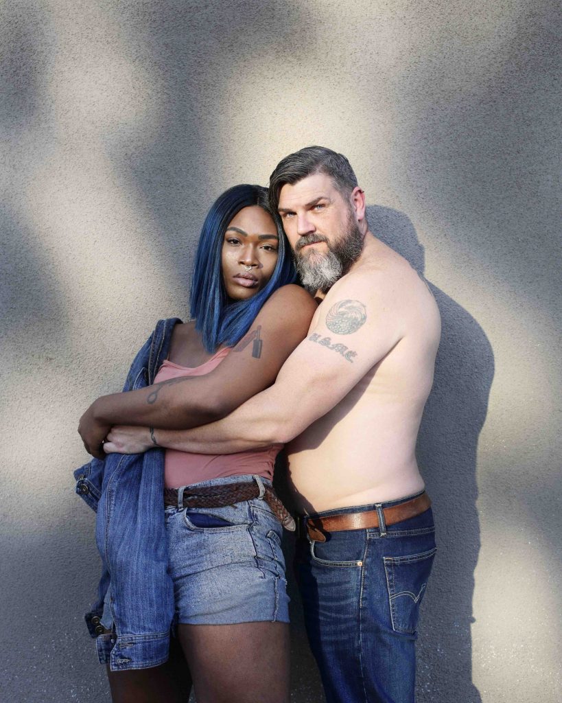 Two people stand, spooning, facing the camera. One is shirtless with a beard and tattoos, while the other has blue and black shoulder length hair and tattoos, and is wearing a singlet and shorts.
