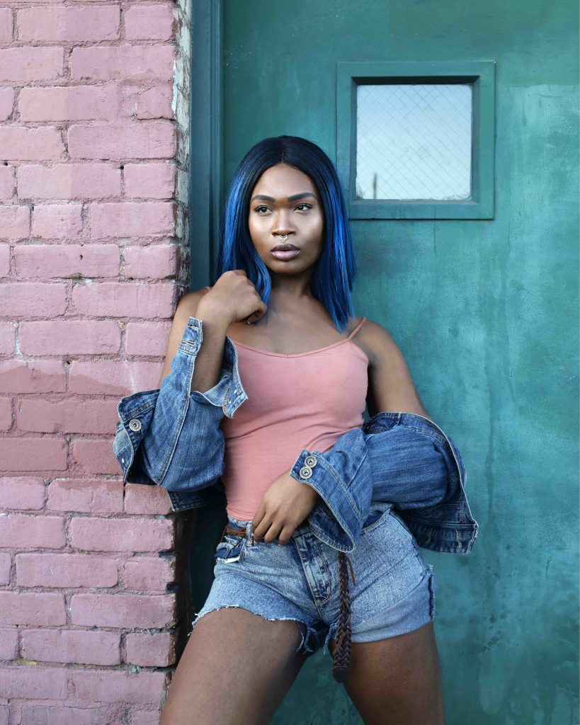 A person stands, facing the camera but looking out of frame. The person has black and blue hair, and is wearing a singlet and shorts with a denim jacket around their arms. 