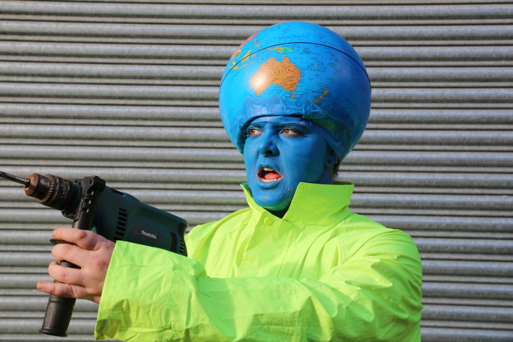 A person with a globe on their head and a blue face to match the colour of the ocean wears a high vis jacket and holds a drill