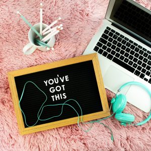 A laptop, turquoise headphones, a sign saying 'you've got this' and a cup of pens