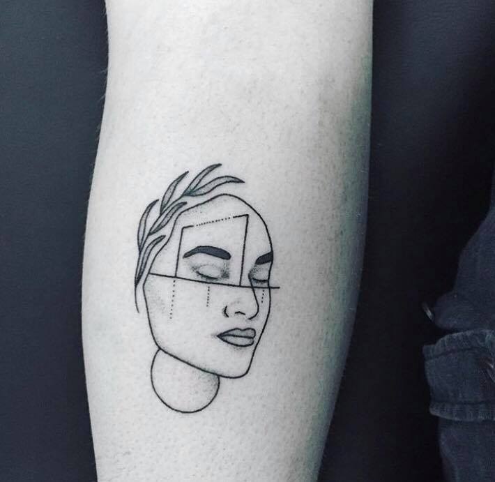 A tattoo of a face with some leaves on top and some geometric lines through the face. The eyes are closed. The image is black and white. 