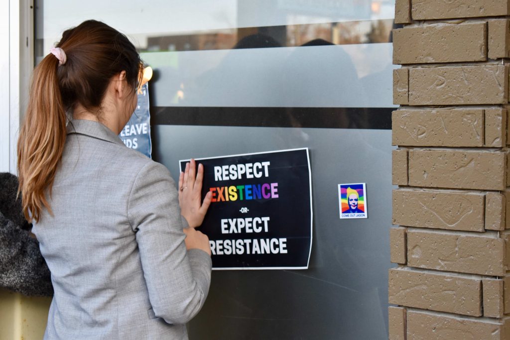 Person with long hair wearing a blazer places a leaflet on the fridge. The leaflet says "respect existence (in rainbow font) or expect resistance."