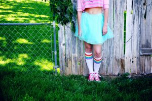 Portrait of a young person standing against a wooden fence wearing rainbow, blue and pink for Pride.