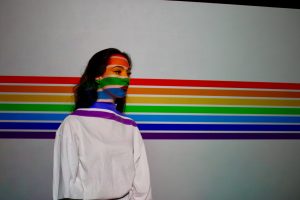 Person standing in front of a white wall with rainbow stripes running horizontally across them and the wall