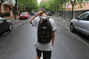 a young adult stands with their back to the camera wearing a backpack, running a hand through their hair.