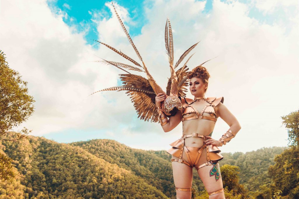 Alyssa wears a bronze coloured costume of harnesses, straps and buckles, one hand on her hip, the other holding an elaborate head dress. She stands in front of a mountain and a blue sky. 