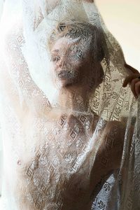 Alyssa faces the camera, visible from the waist up. A piece of lace covers her face and body, but her features are still visible.