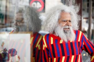Jack Charles wears a colourful jumper, and looks to the right of the frame