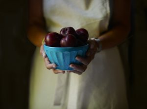 A person in a yellow apron holds a bowl of plums.