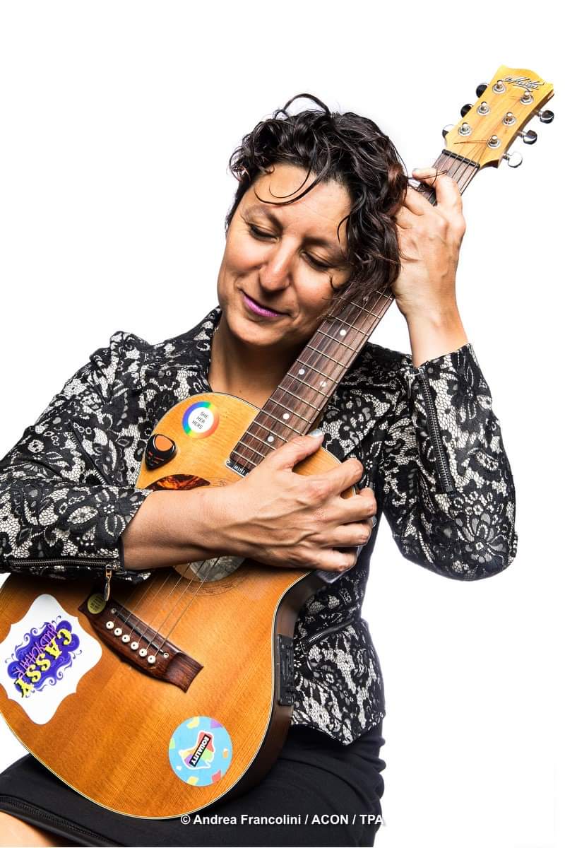 A woman holds a guitar into her chest and smiles with eyes closed