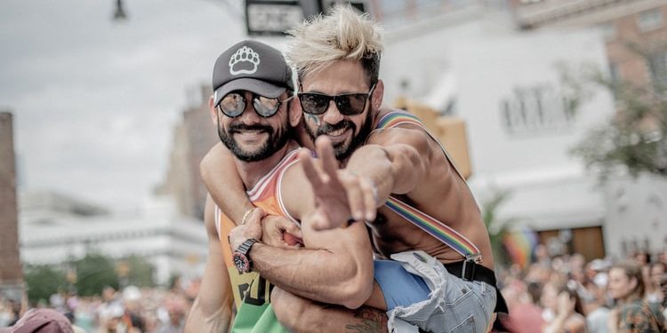 One man holding onto another mans back, making peace sign at camera and smiling