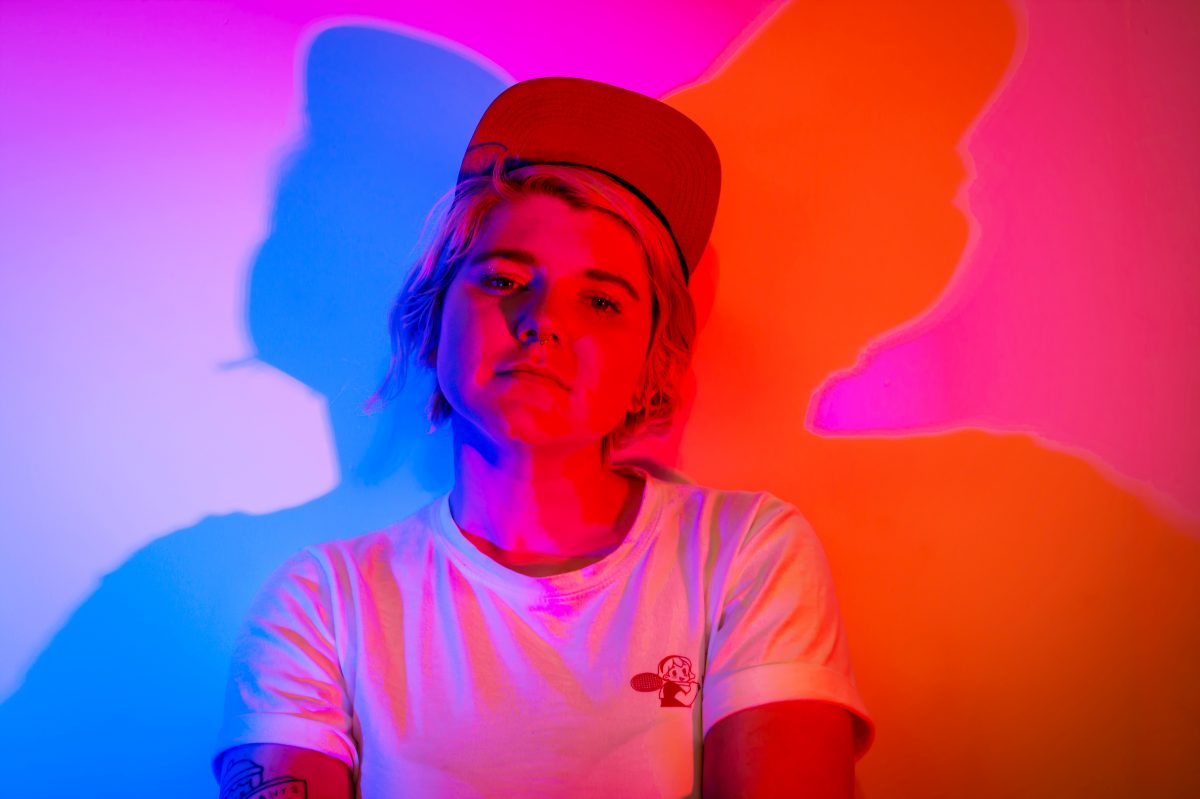 Photo of the author Dani Leever, a white non binary person wearing a hat, a white shirt and they have short hair. The lighting is dark pink and blue.