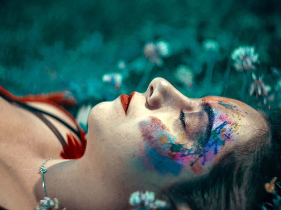 Person lying on the ground with their eyes closed and wearing colourful paint on their face