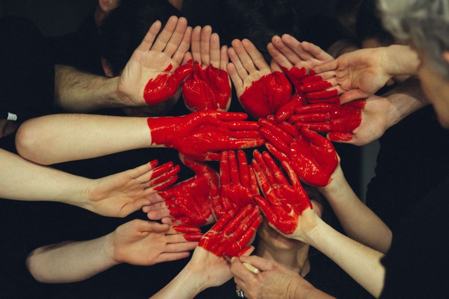 Multiple hands, palms facing up and covered in bright red paint to varying degrees, are placed together to form a heart.
