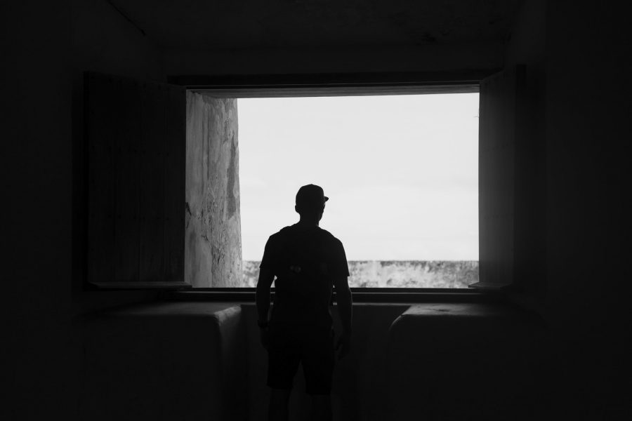 A silhouette of a person wearing a cap stands in front of a glassless window in a very dark room, staring out into the light. The image is black and white.