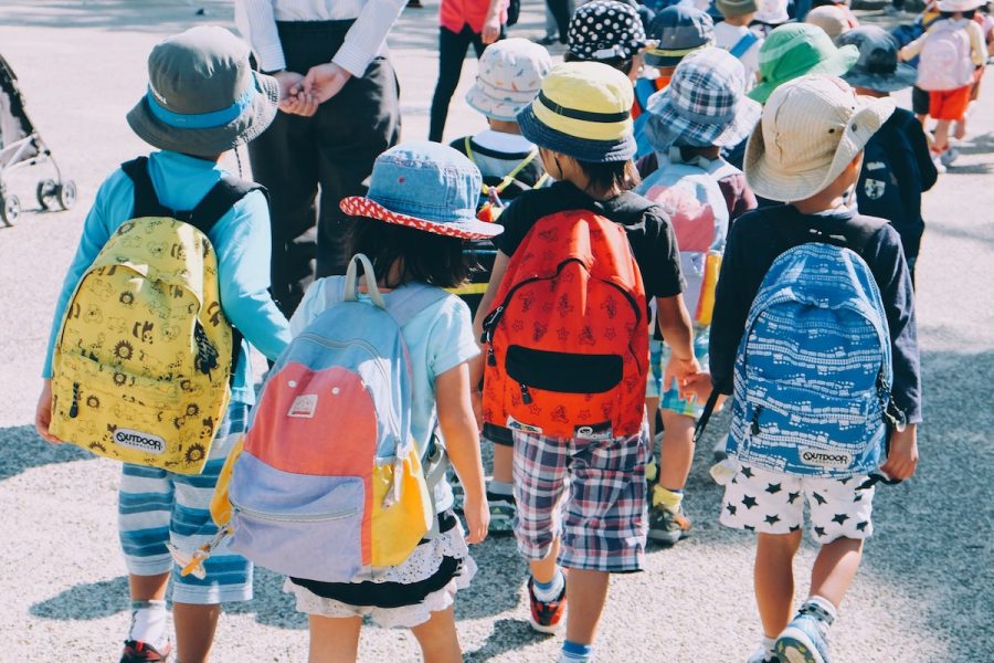 A photo of children all walking away from the camera wearing colourful backpacks. There is a large group of children, with four that are closest to the camera.