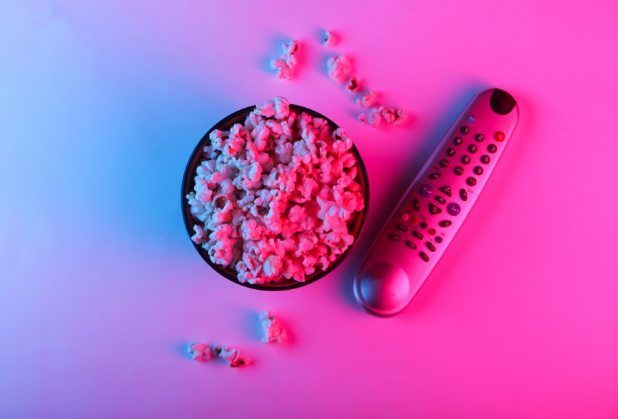 A TV remote and bowl of popcorn are seen from above, bathed in neon pink blue light.