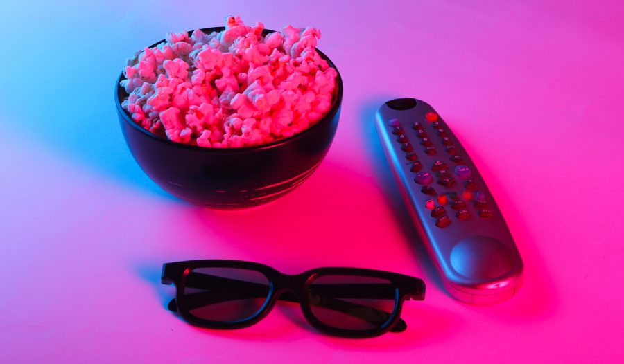 A TV remote, a pair of glasses, and a full bowl of popcorn on a table are illuminated in deep pink and purple light.