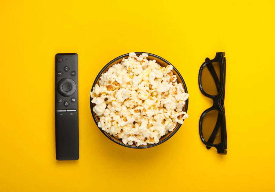 A TV remote, a bowl of popcorn, and a pair of sunglasses are seen from above against a bright yellow background