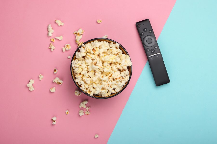 A bowl of popcorn and a slim, black TV remote are viewed from above. The background is split in two diagonally, slightly left of centre. The left hand side is pink pastel, and the right hand side is blue pastel.