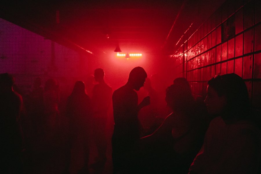 A dark red room that looks like a club. There are tiled walls and the silhouettes of numerous people are seen in the frame.