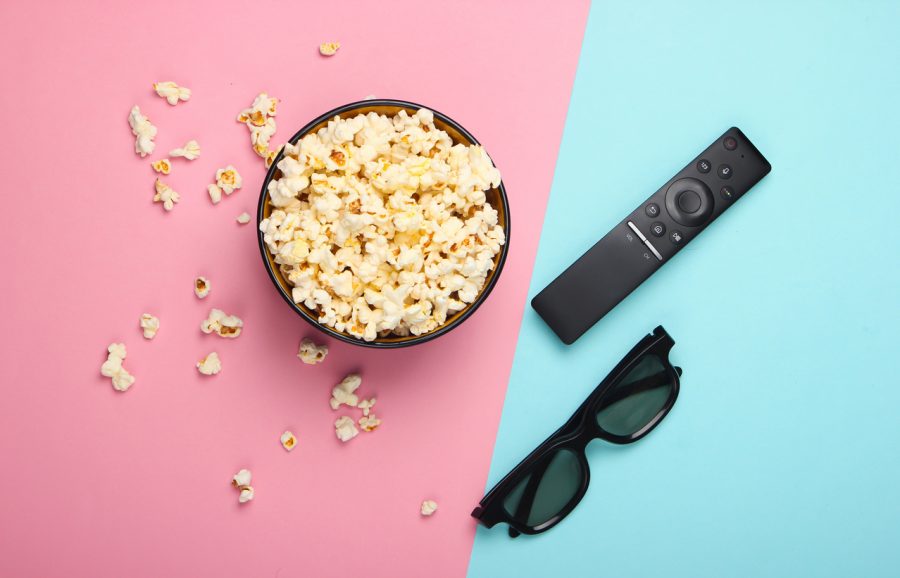 A bowl of popcorn, a pair of glasses, and a slim, black TV remote are viewed from above. The background is split in two diagonally, slightly left of centre. The left hand side is pink pastel, and the right hand side is blue pastel.