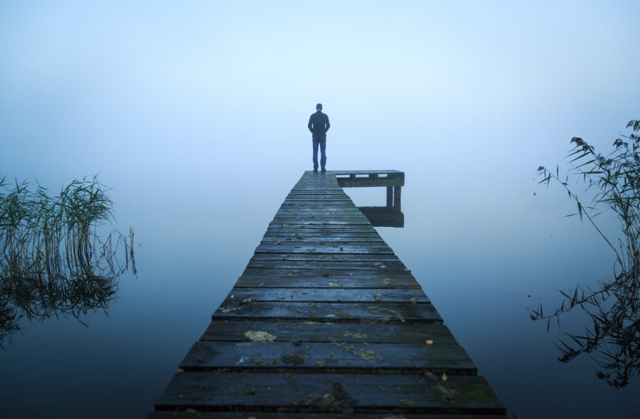 A figure stands alone at the end of a long jetty on a foggy, overcast day. They are looking out over the water which is grey blue and blends into the sky.