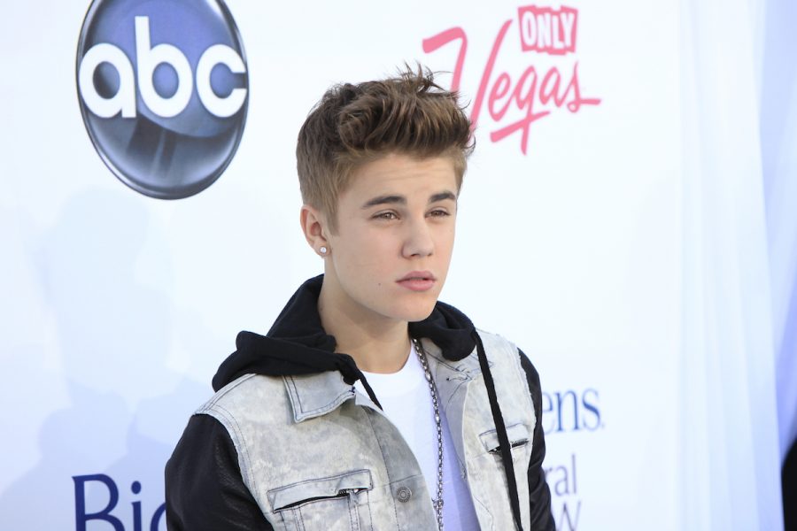 Justin Bieber, a white young man, stands on a red carpet in a close up photo. He is wearing a denim vest with a hood, has short brown styled up hair and is looking smouldering toward the right of the camera.
