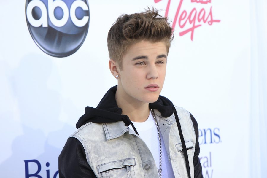 Justin Bieber, a white young man, stands on a red carpet in a close up photo. He is wearing a denim vest with a hood, has short brown styled up hair and is looking smouldering toward the right of the camera.