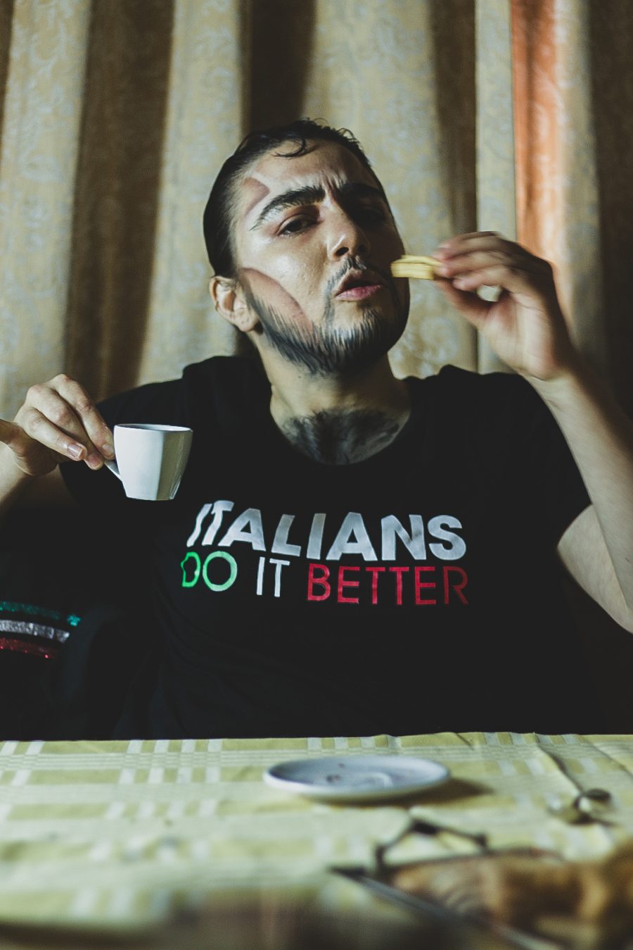 The author in drag as Bruno Salsicce. They're a drag king sitting at a table holding a small espresso cup and eating a biscuit. They have slicked hair, chest and facial hair, drag king makeup and their shirt says "Italians do it better"